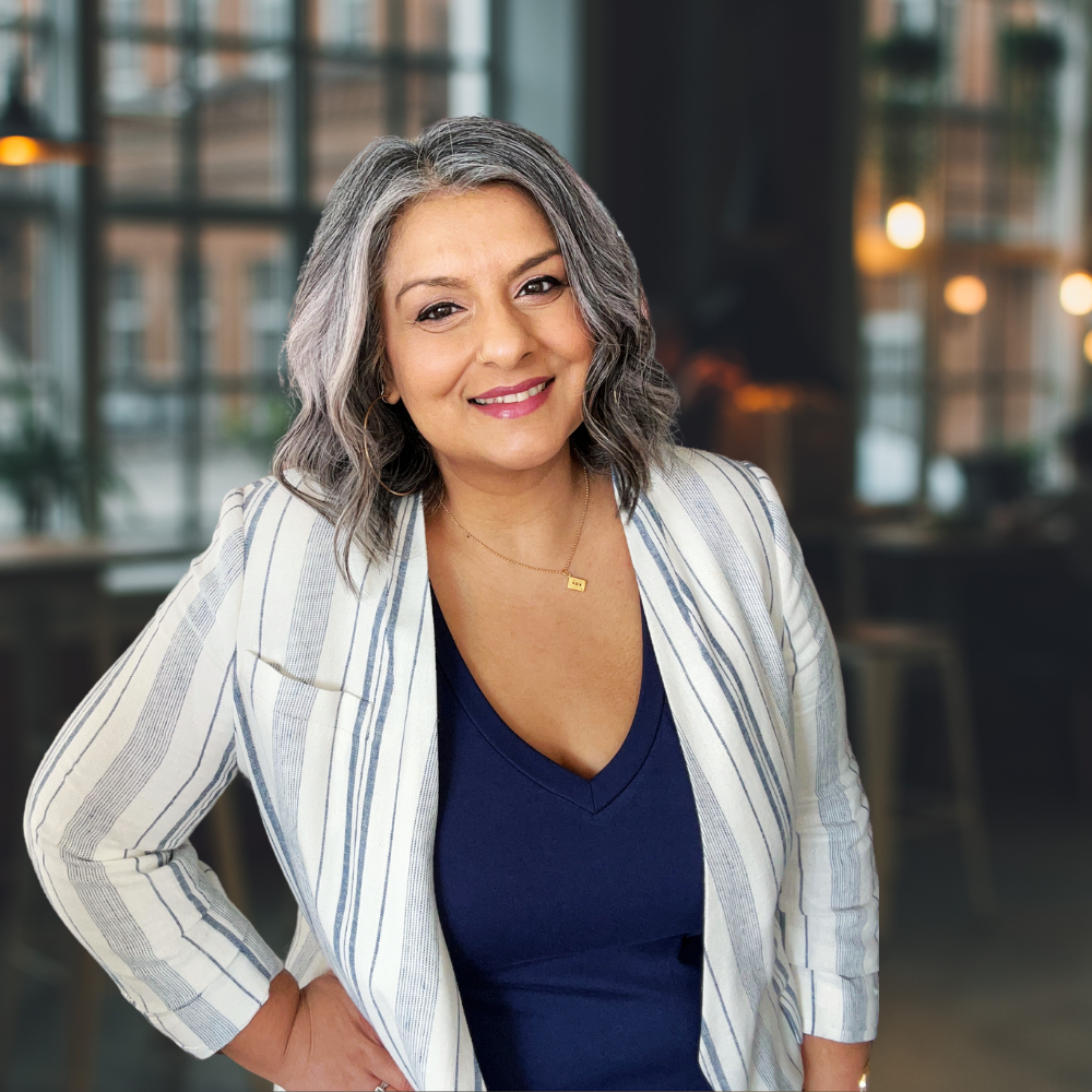 Puja McClymont headshot she wears a blue and white jacket with blue vest smiling at camera whilst leaning head to one side